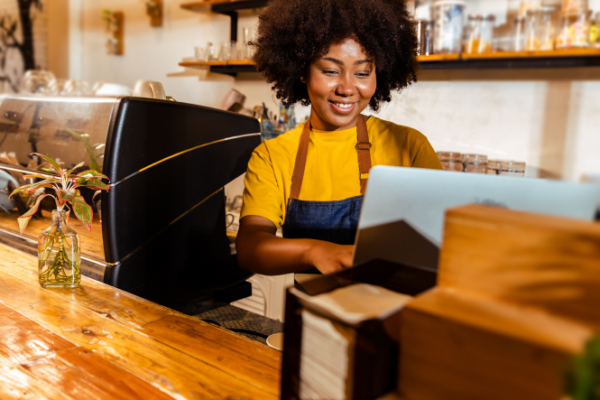 A barista works on a laptop behind the counter of a coffee shop.