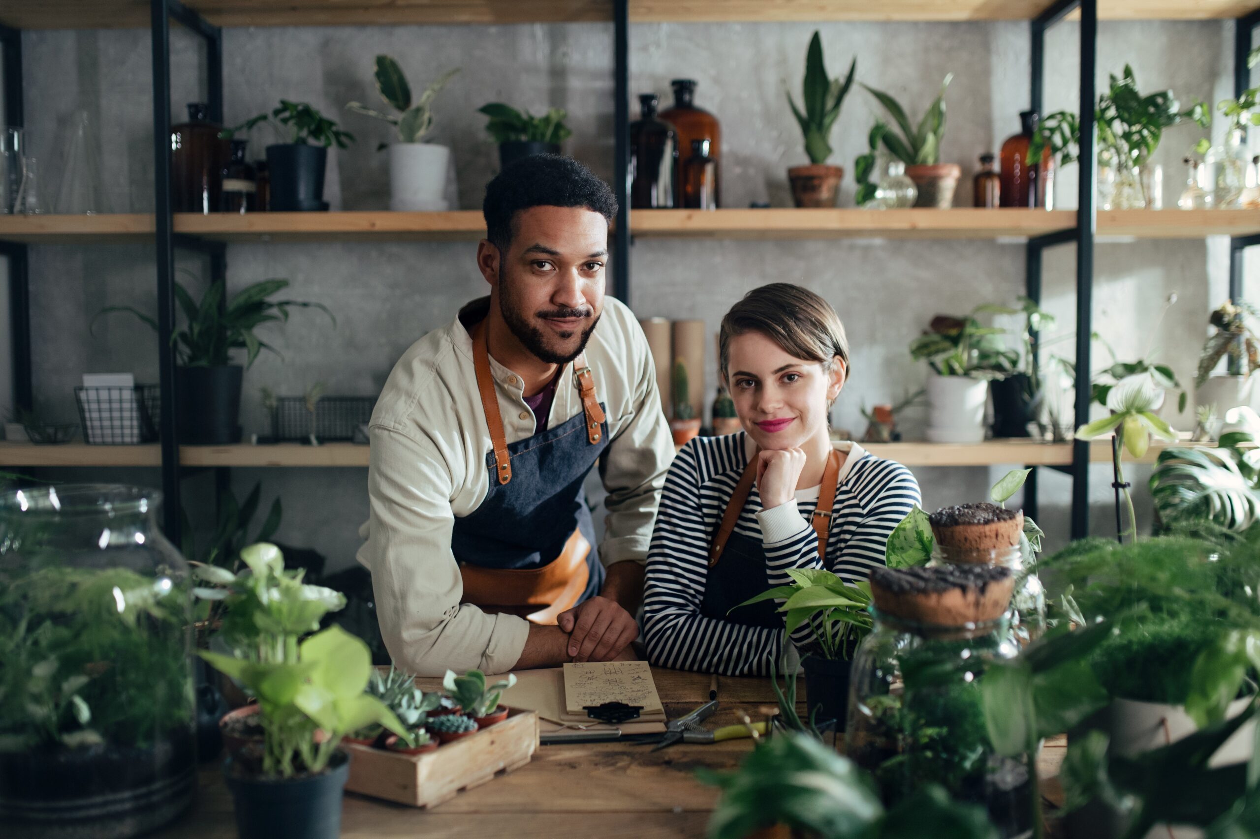 An image of a man and woman in aprons leaning on a counter with various plants and suculents around them.
