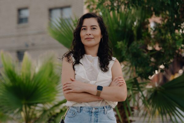 A woman stands proudly with her arms crossed in front of a building and palm trees.