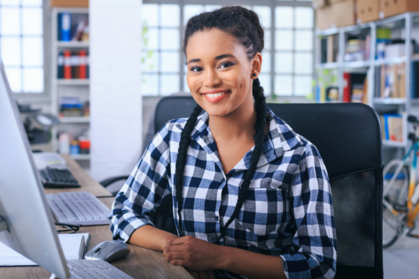 Smiling young woman sitting at a desk in a modern office, wearing a plaid shirt and working on a desktop computer