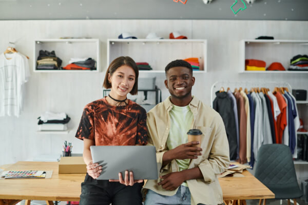 Two smiling designers in a clothing store, holding a laptop and coffee cup.